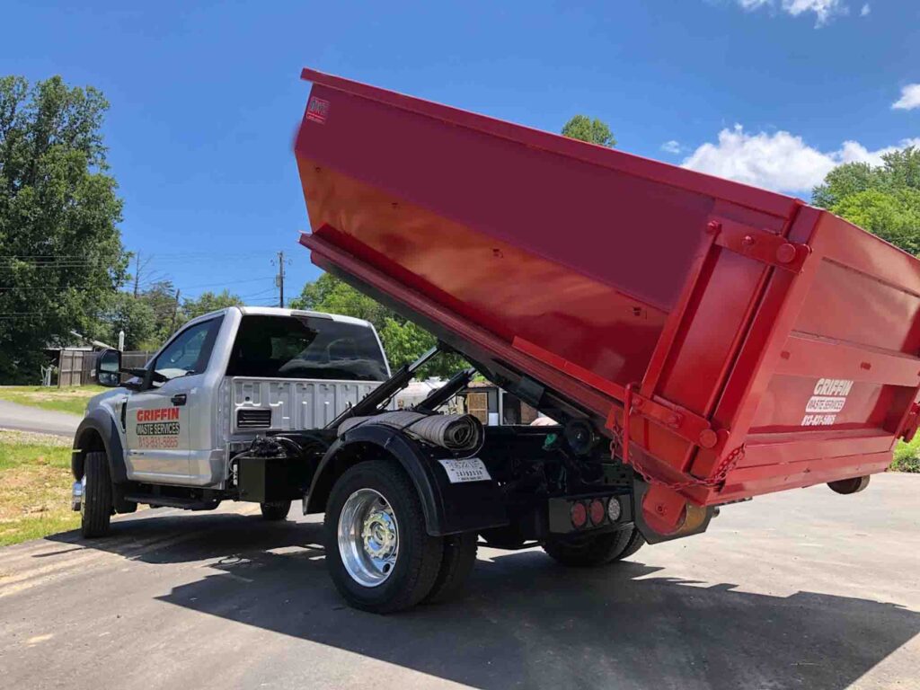 A gray and black truck with a red dump bed raised in a tilted position, parked on a paved surface. Griffin Waste Services is visible on the side of the truck. Trees and a blue sky in the background.