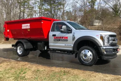A white Griffin Waste Services truck with a large red dumpster container on its bed is parked on wet pavement, offering convenient dumpster rental in Lake Keowee and Hartwell, with trees and grass in the background.