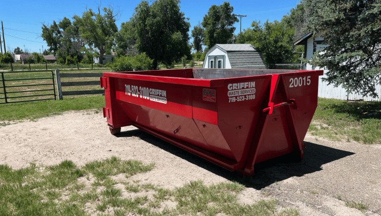 A large red dumpster labeled GRIFFIN Waste Services, perfect for dumpster rental Colorado Springs, sits on a patch of dirt and grass near a white shed and trees in a rural area under a sunny sky.