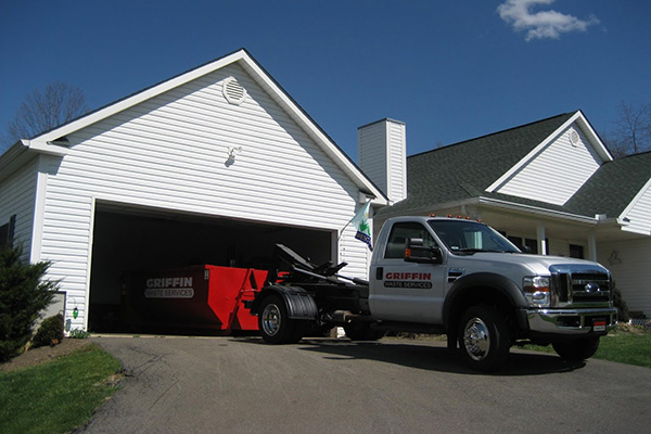 A white truck with Griffin Waste Services branding is parked in the driveway of a white house with a green roof. The truck is attached to a red dumpster, partially visible inside the garage. The sky is clear with a few clouds.