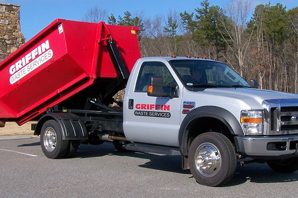 A silver and red waste disposal truck is parked on a road. The truck, labeled Griffin Waste Services, has a large red container on its back. The background features trees and a clear sky.