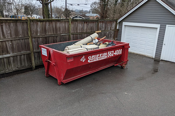 A red dumpster labeled Griffin Waste Service beside a wooden fence and a gray garage. Inside the dumpster are various rolled-up carpets and some construction debris. The scene is on a paved driveway with overcast weather.