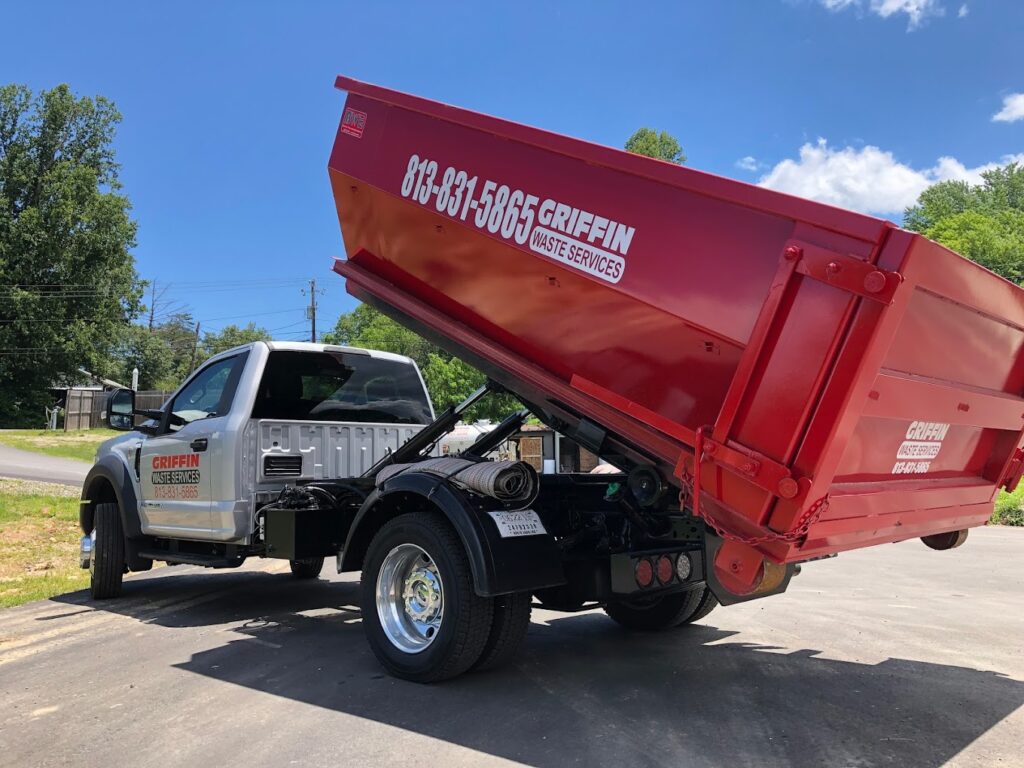 A white truck with a red dumpster rental container in a lifted position is parked on a paved surface. Green trees and a clear blue sky serve as the backdrop, while the container prominently displays the phone number and name of a waste services company.