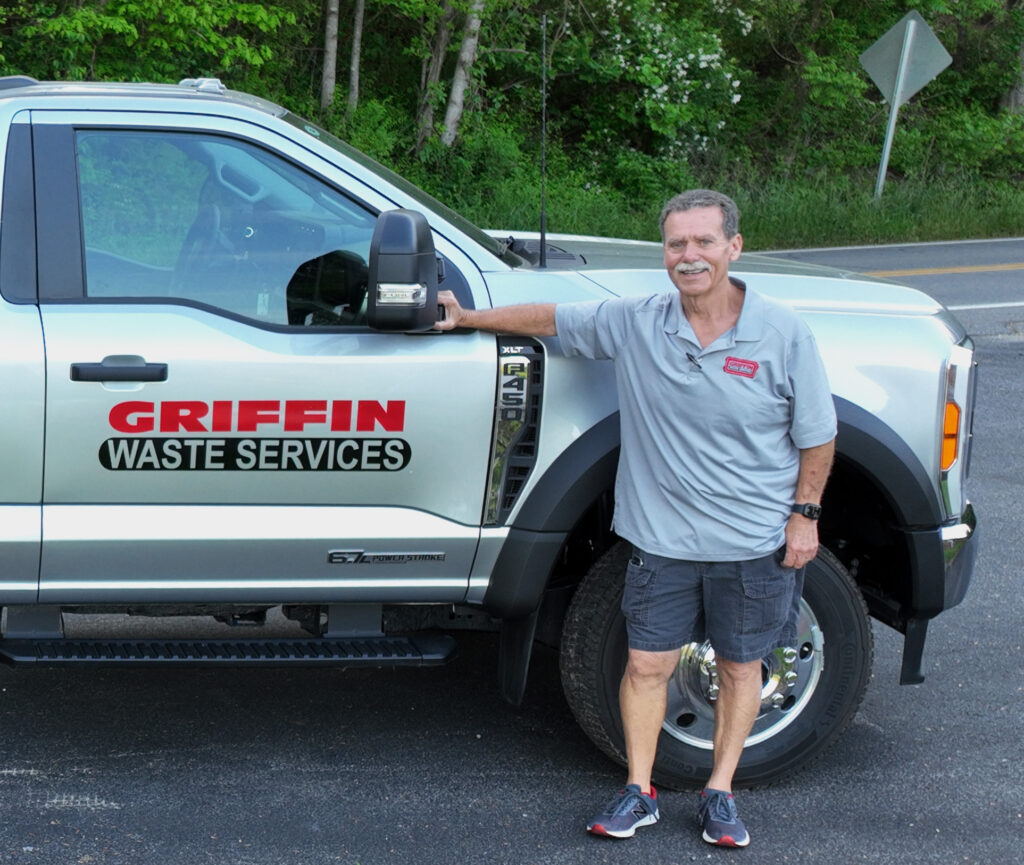 A man in a gray shirt stands smiling next to a silver truck with Griffin Waste Services written on the door. He is resting his arm on the vehicle. The background features green foliage and a road.