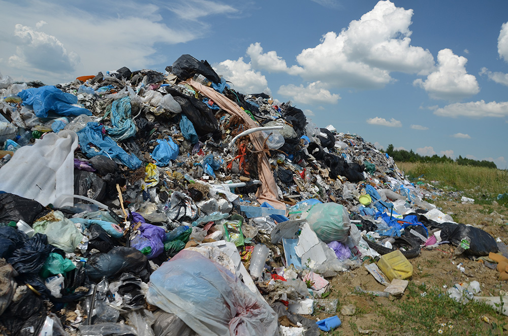 A large pile of mixed garbage, including plastic bags and various waste materials, sits on open ground under a partly cloudy sky, with some grass and trees visible in the background.