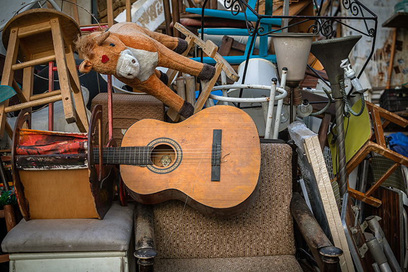 A cluttered pile of assorted objects including a worn acoustic guitar, a brown armchair, a toy horse, lamps, stools, and various household items stacked haphazardly together.