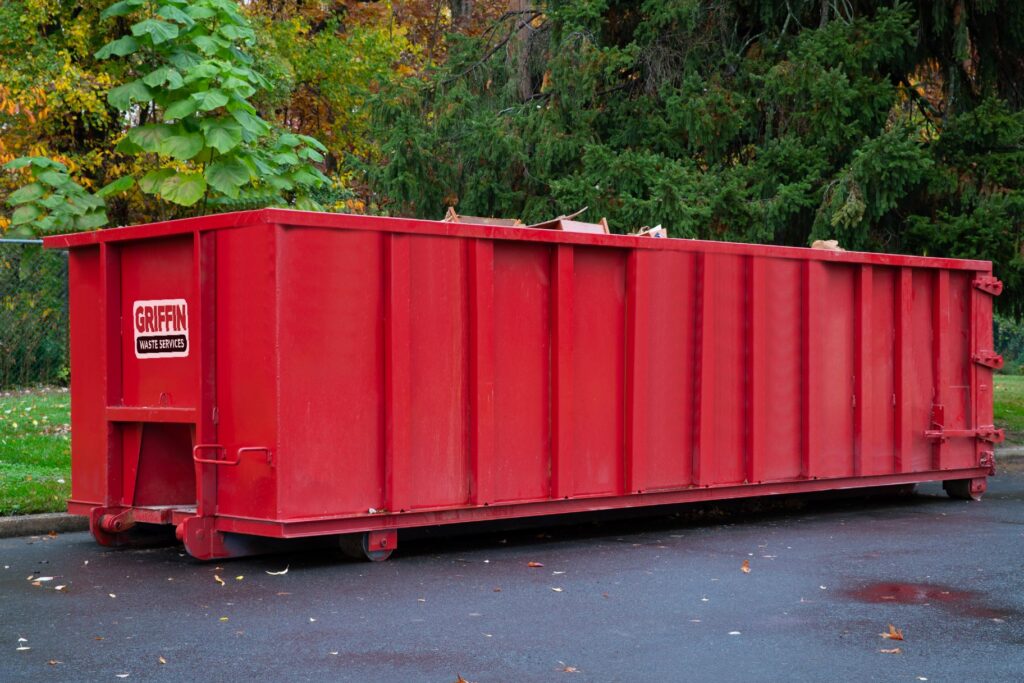A large red dumpster labeled Griffin Waste Services sits on a paved surface near green grass and trees with autumn foliage in the background.