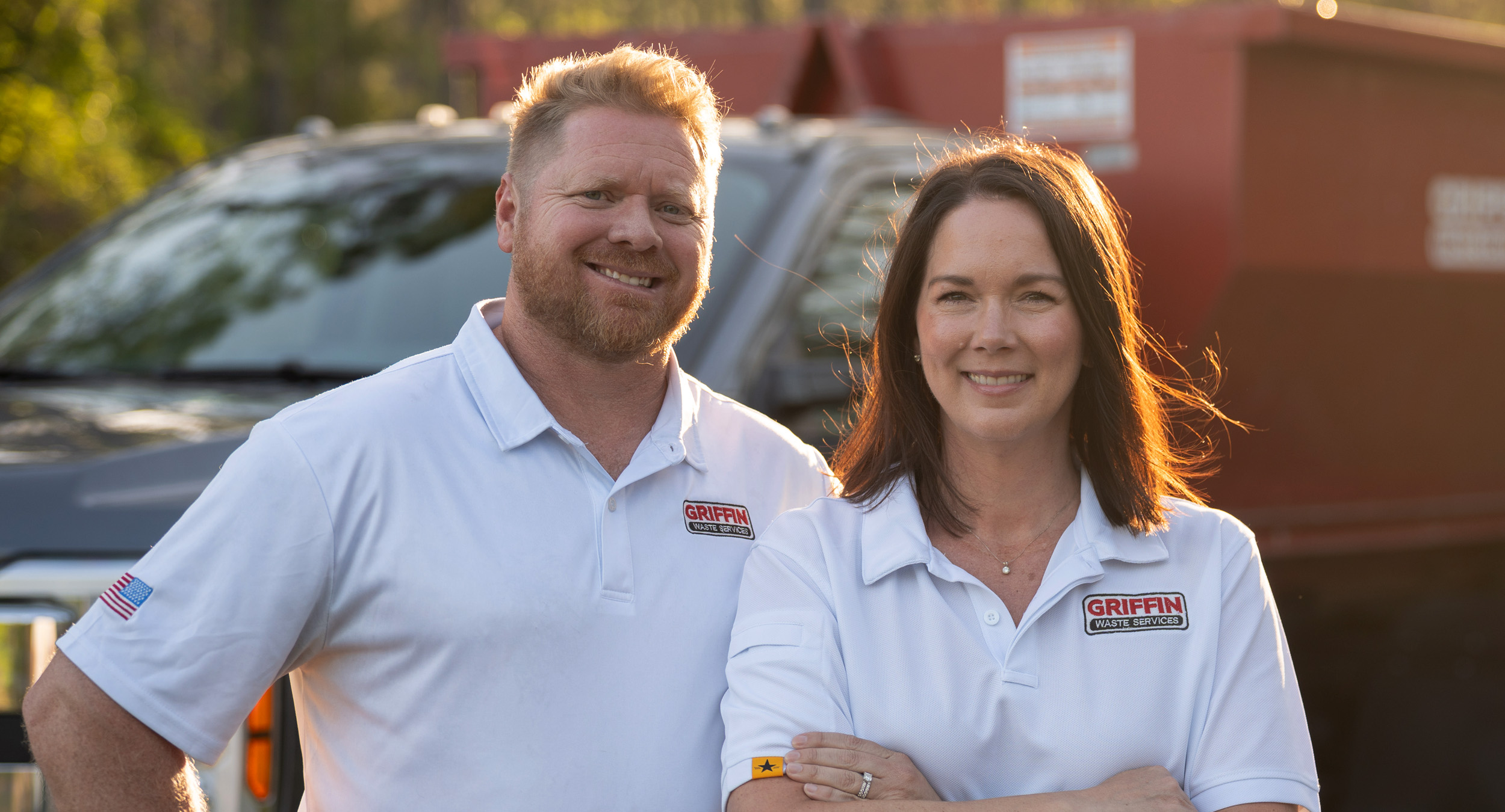 griffin_waste_owners A man and a woman wearing white polo shirts with Griffin logos stand outside in front of a black truck and a red container, smiling at the camera.