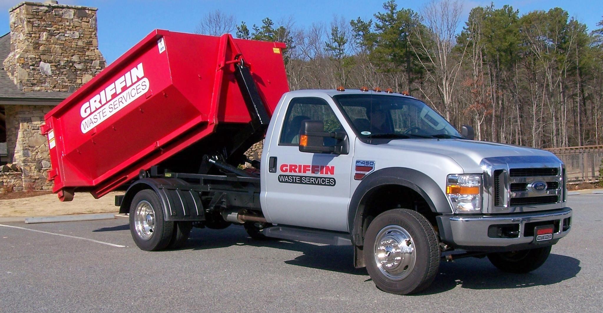 A delivery truck is backing a roll-off dumpster down a residential street, which is lined with mature trees. This scene illustrates the reliable service of dumpster rental in Knoxville, perfect for yard waste, home renovations, or construction projects.