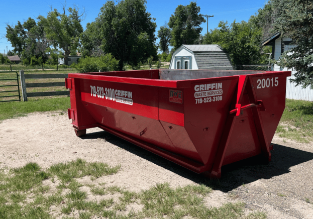 A large red dumpster labeled GRIFFIN Waste Services, perfect for dumpster rental Colorado Springs, sits on a patch of dirt and grass near a white shed and trees in a rural area under a sunny sky.