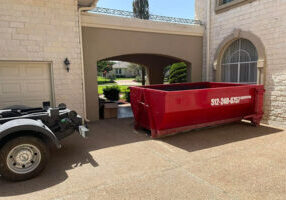 A red dumpster with a phone number is placed in a driveway near a garage and arched entrance of a light-colored brick house. The rear part of a truck or vehicle is visible, suggesting delivery or placement of the dumpster.