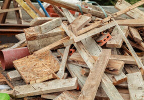 A pile of assorted wooden planks and construction debris, including pieces of wood, nails, and a few bricks, scattered outdoors at what appears to be a construction or demolition site.