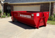A large red dumpster is situated on a concrete driveway in front of a brick house with a two-car garage. The dumpster displays the number 940-326-3047 and the words Griffin Waste Services.