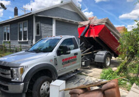 A gray truck with Griffin Waste Services on the side is parked in a driveway, unloading a large red dumpster. A blue house and some outdoor furniture are visible nearby under a partly cloudy sky.