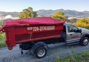 A red and silver waste services truck is parked on a dirt road with mountains and trees in the background. The truck has a red cover over its cargo area and contact information on the side.