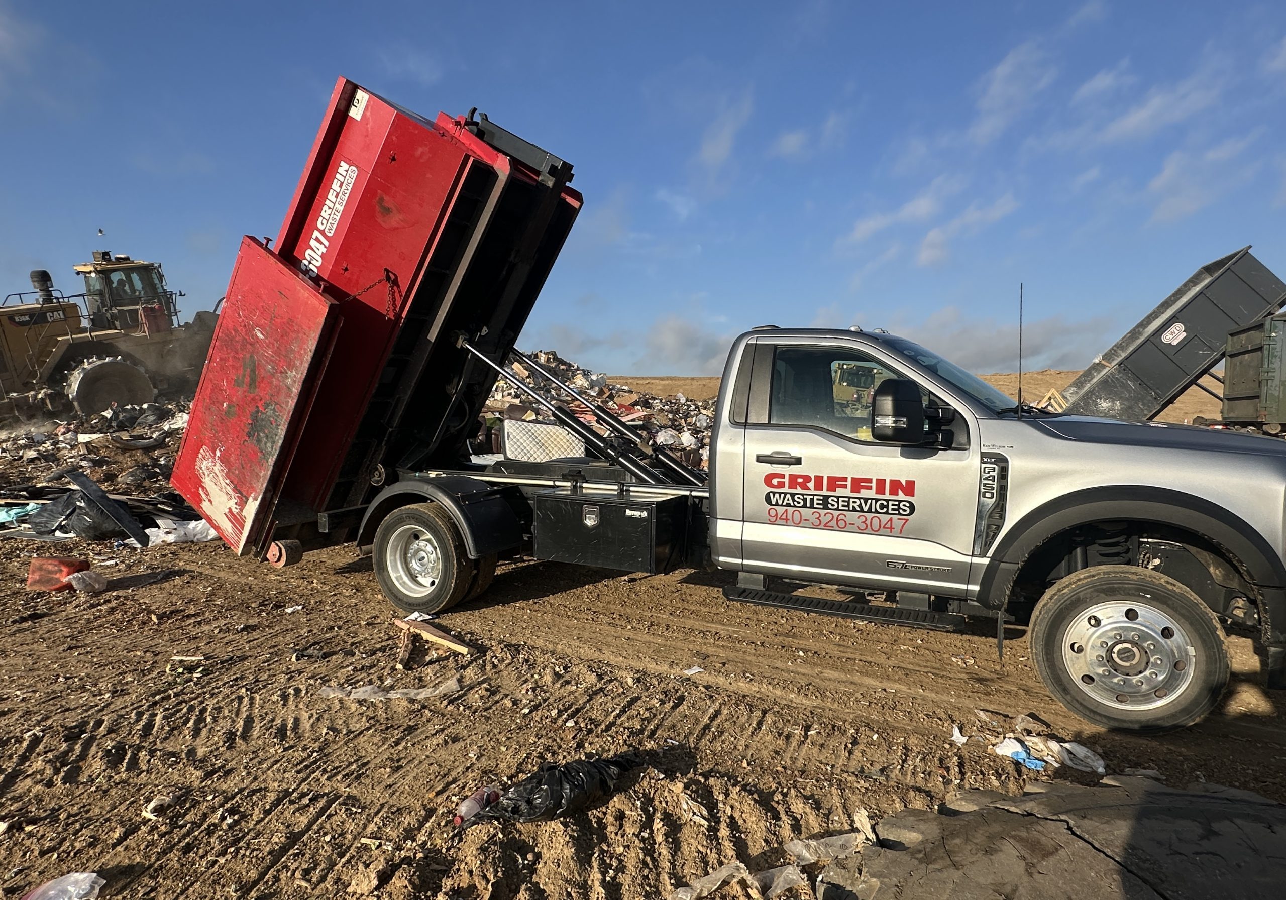 A Griffin Waste Services truck unloads a red dumpster at a landfill, surrounded by piles of trash and dirt, under a partly cloudy sky.