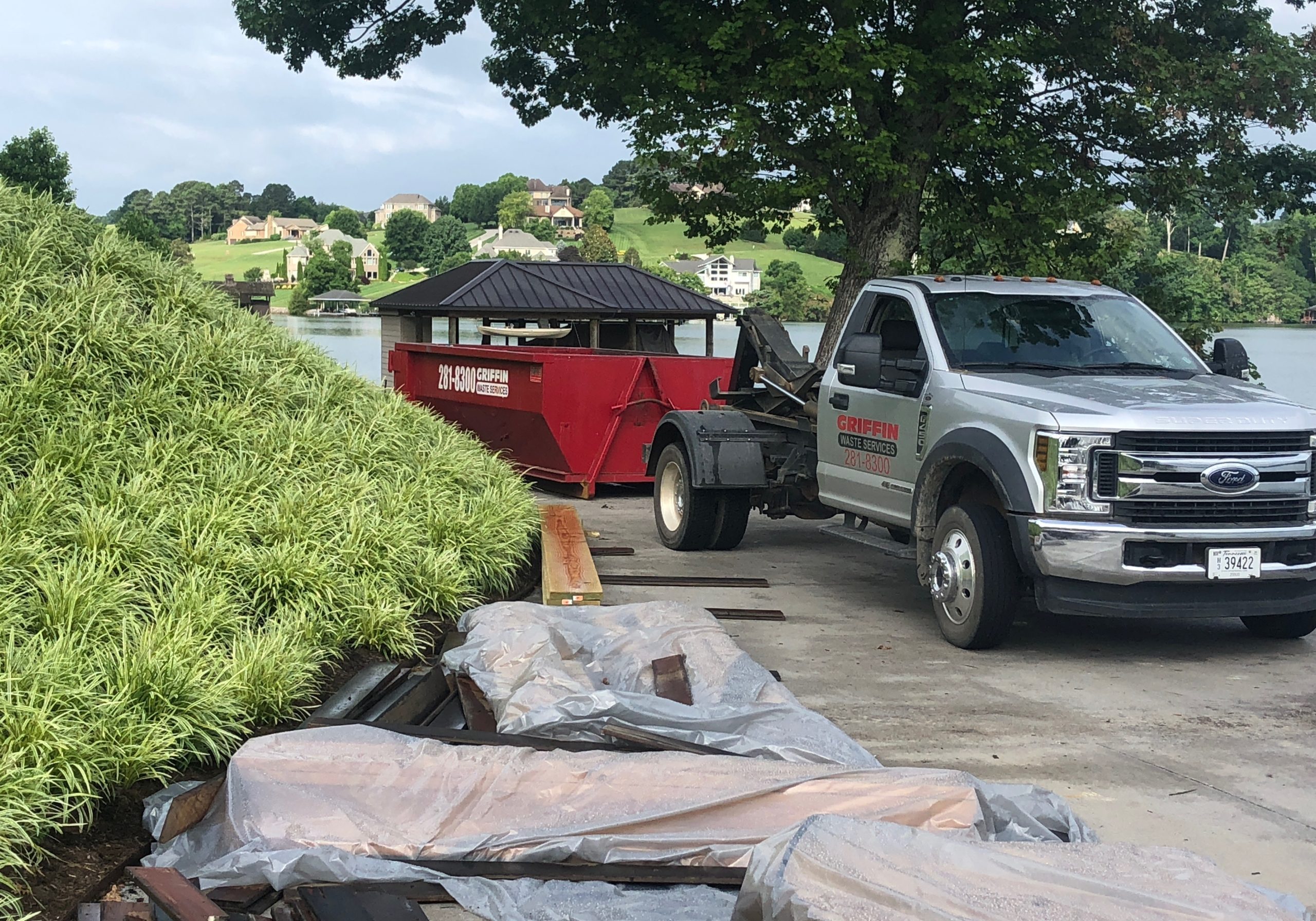 A GreerBin dumpster truck is parked on a lakeside driveway near a red dumpster, with construction materials and covered items in the foreground and homes visible across the water.