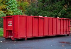 A large red dumpster labeled Griffin Waste Services sits on a paved surface near green grass and trees with autumn foliage in the background.