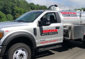 A silver waste service truck, featuring Griffin Waste Services Asheville NC on its side, is parked along the road. Known for its portable toilet rental services, the truck sits against a backdrop of trees and a clear sky.