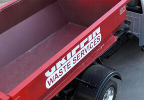 A red waste services truck with an open container, showing the words Waste Services in white. The truck is parked on a gray surface, and part of the cab is visible on the right.