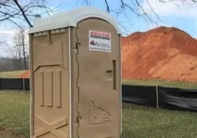 A beige portable toilet rental unit stands on grass near a construction site. Behind it, theres a large pile of orange soil and a black mesh fence. Trees without leaves are visible in the background under a clear blue sky.