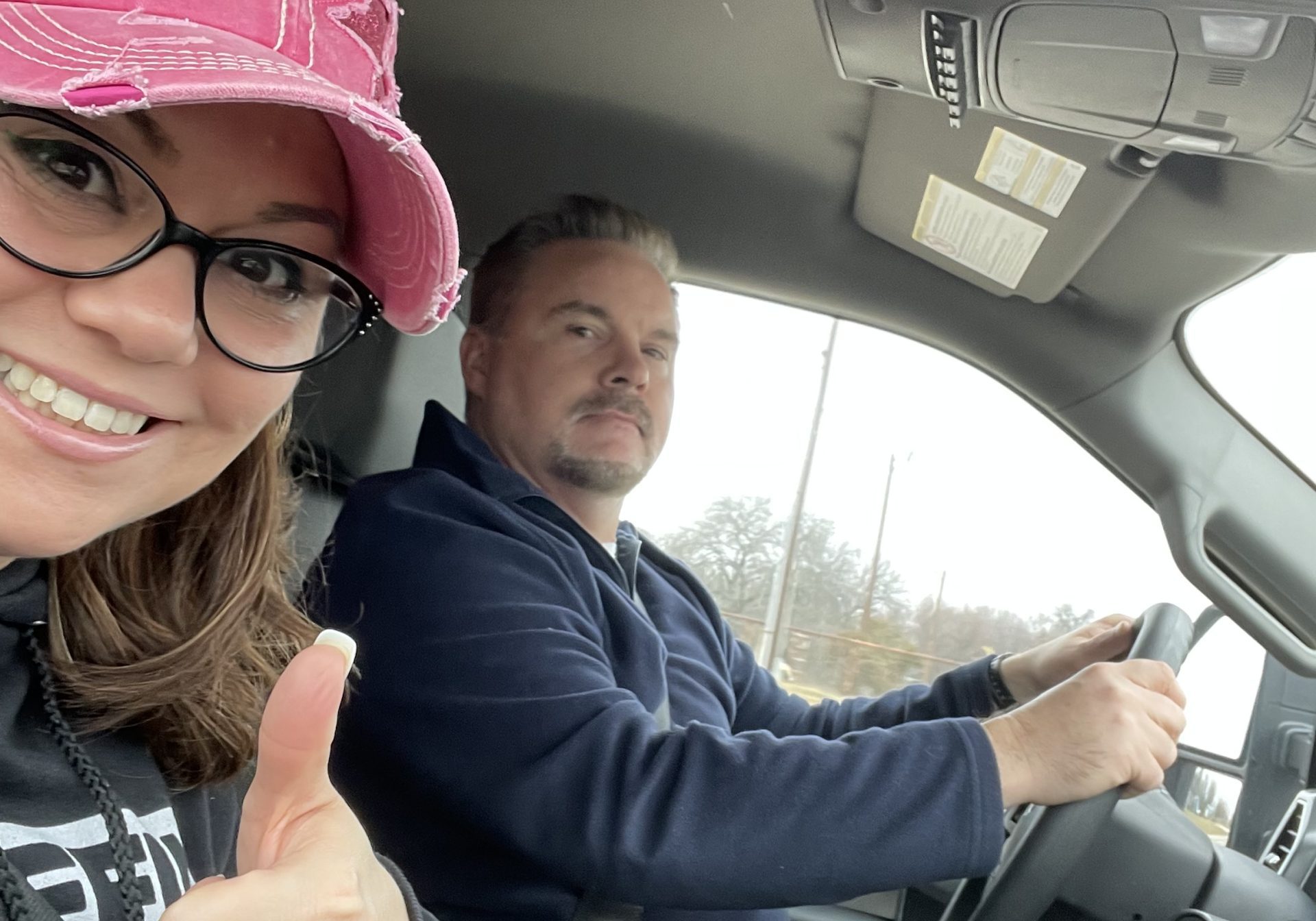 A woman wearing a pink cap and glasses gives a thumbs-up and smiles at the camera while sitting beside a man driving a car. The man looks forward with a neutral expression. A cup and red cord are on the console.